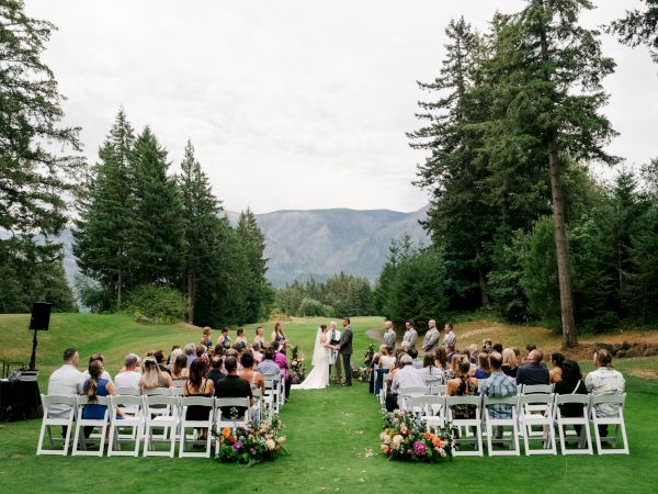 An outdoor wedding ceremony with guests seated on a lawn, surrounded by trees and mountains in the background.