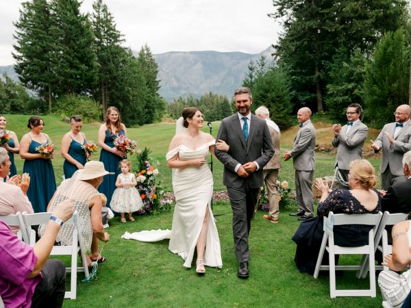 A couple walks arm in arm down an outdoor aisle, surrounded by happy guests and a wedding party, while people applaud in celebration.