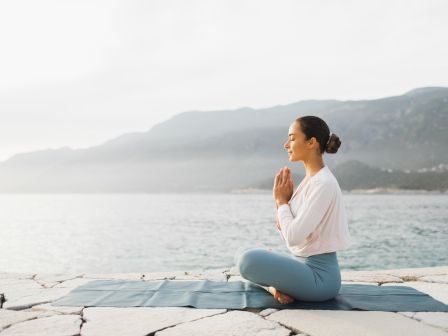 A person is meditating in a seated position on a yoga mat by the water, with mountains in the background.