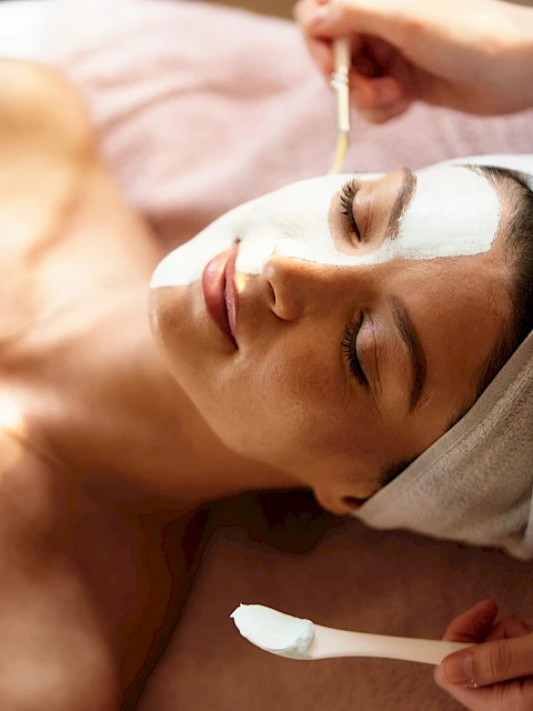 A woman is receiving a facial treatment, with cream applied to her forehead and eyes, lying down with a towel around her hair.