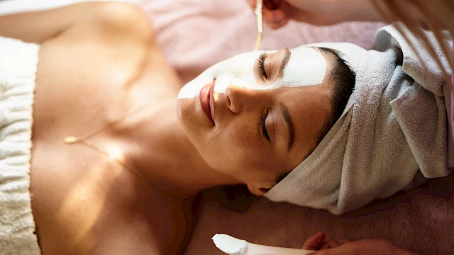 A woman is receiving a facial treatment, with cream applied to her forehead and eyes, lying down with a towel around her hair.