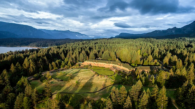 A large building surrounded by dense forest, mountains, and a body of water under a cloudy sky.
