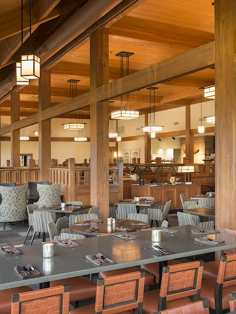 The image shows a spacious, modern restaurant interior with wooden beams, neatly arranged tables and chairs, and soft overhead lighting.