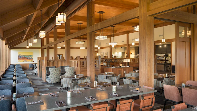 The image shows a spacious, modern restaurant interior with wooden beams, neatly arranged tables and chairs, and soft overhead lighting.