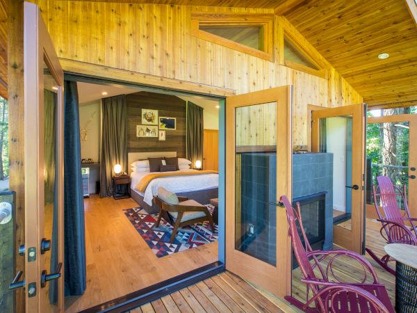 A wooden cabin doorway opens to a cozy bedroom with a bed, rug, and lamp, plus an outdoor deck with purple chairs and forest scenery.