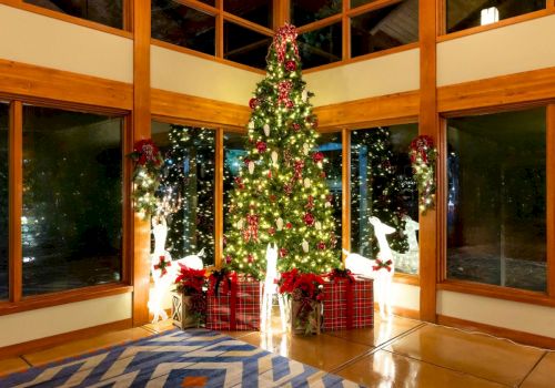 A decorated Christmas tree with red ornaments stands in a bright room, surrounded by gift boxes near large windows.
