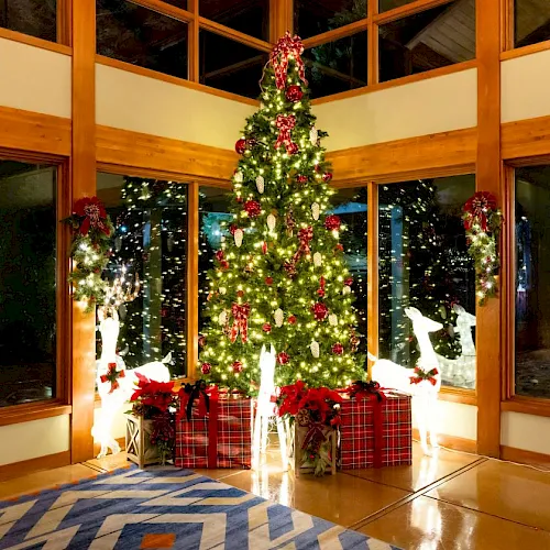 A decorated Christmas tree with red ornaments stands in a bright room, surrounded by gift boxes near large windows.