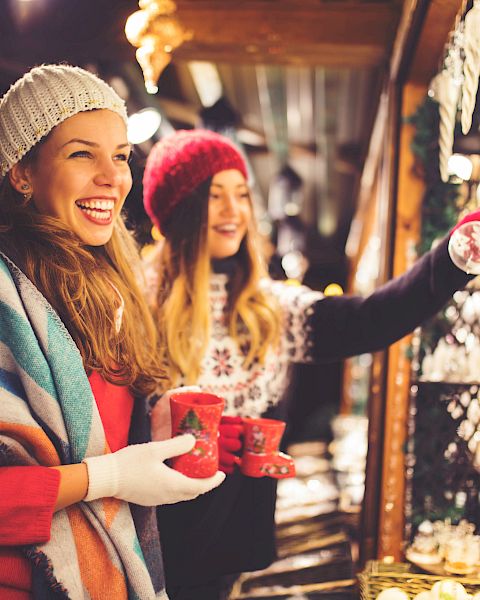 Two women in winter attire, with mugs in hand, enjoying a festive market surrounded by lights and holiday decorations.