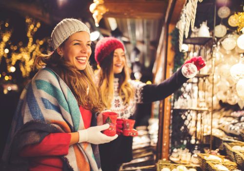 Two women in winter attire, with mugs in hand, enjoying a festive market surrounded by lights and holiday decorations.