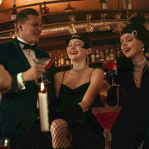 A 1920s-themed party in a bar: women and men in formal attire toast with cocktails and laugh under warm lights.