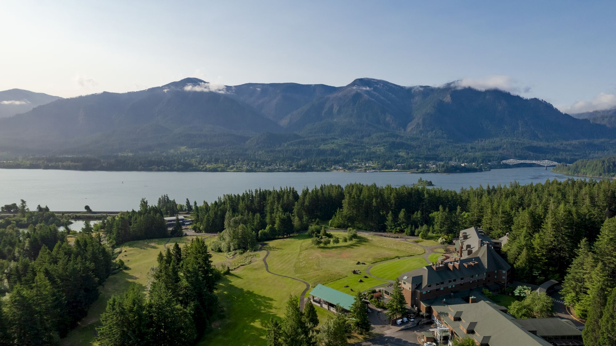A scenic landscape with a lake, distant mountains, and a large building surrounded by trees and open greens in the foreground.