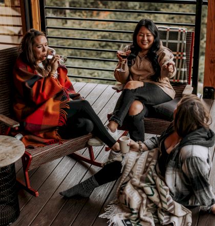 Three people are relaxing on a deck with drinks, surrounded by nature.