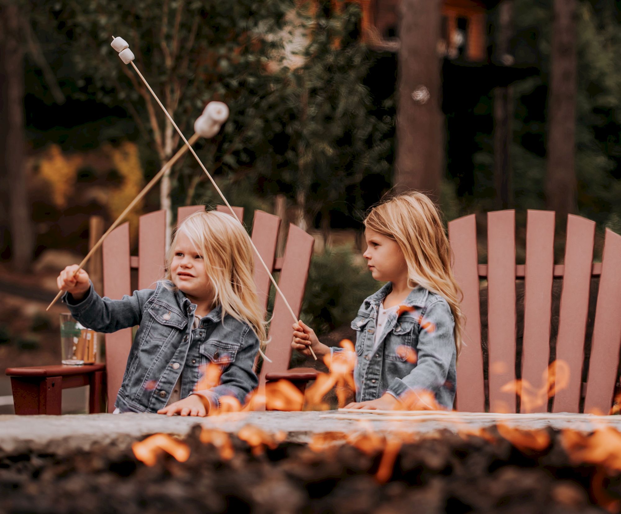 Two young girls sit by a fire pit in denim jackets, roasting marshmallows on long sticks.