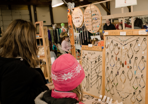People are browsing jewelry at a market stall. The display includes necklaces and bracelets on a wooden stand, indoors.