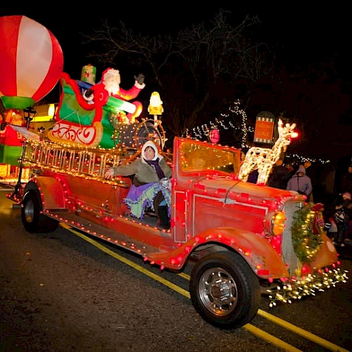 A festive, brightly lit parade float with Christmas decorations, including a reindeer, Santa, and a large balloon on a classic red vehicle.