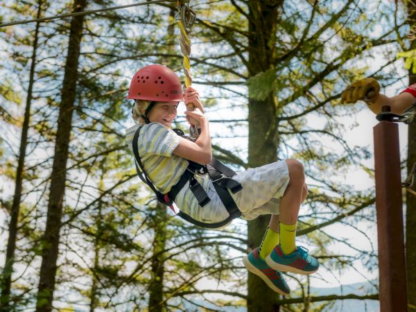 A child wearing a helmet and harness enjoys a zipline ride in a forest setting, with a guide assisting nearby.